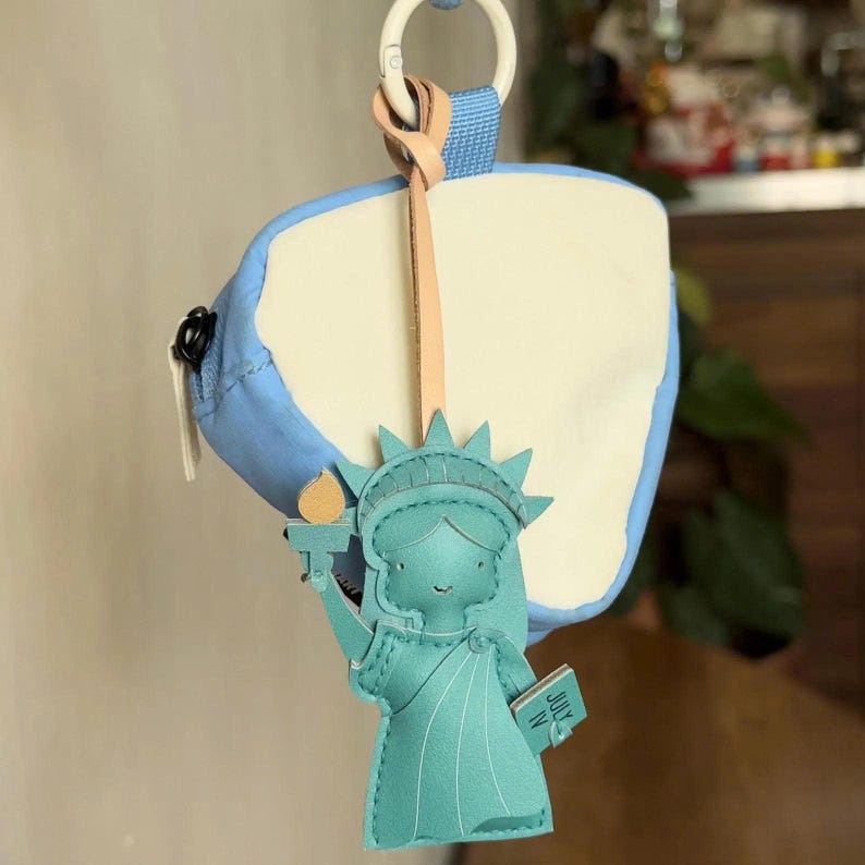 May include: A light blue and cream colored pouch with a white plastic ring attachment. A teal Statue of Liberty charm hangs from a tan leather strap, featuring a yellow torch flame and a book with the text JULY IV printed on the cover.