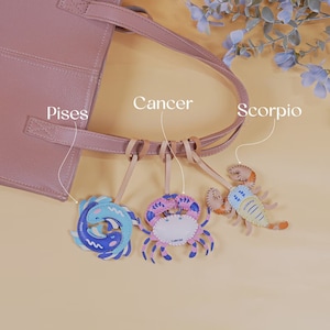Puede incluir: Un bolso tote de cuero rosa con tres dijes de cuero unidos a las asas. Los dijes tienen forma de pez, cangrejo y escorpi&oacute;n. Los dijes est&aacute;n decorados con pintura azul, rosa y amarilla y tienen las palabras "Piscis", "C&aacute;ncer" y "Escorpio" escritas en ellos.