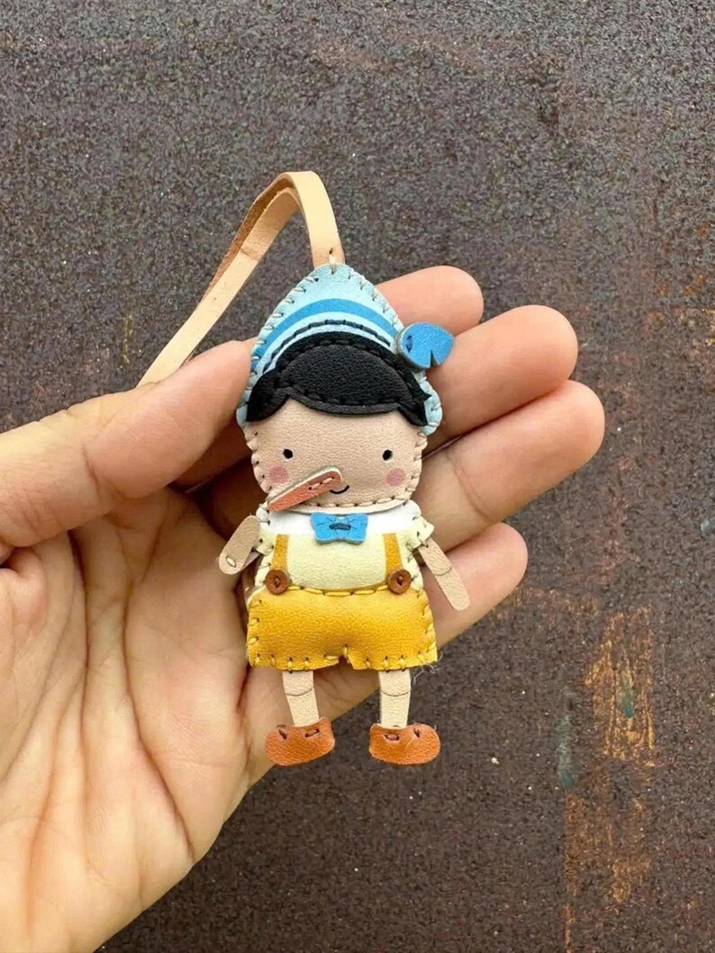 May include: A handmade leather Pinocchio doll with a light blue and white striped hat, black hair, and a long nose. The doll wears a white shirt, yellow overalls, and brown shoes. A tan leather loop is attached to the top of the doll.