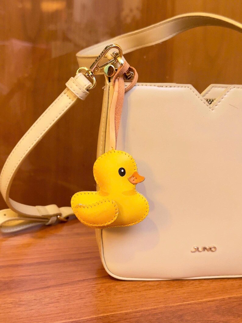 May include: A cream-colored handbag with a yellow rubber duck charm attached. The bag has a shoulder strap and a gold-colored clasp. The duck charm is yellow with an orange beak and black eyes. The word "JUNO" is printed on the bag.
