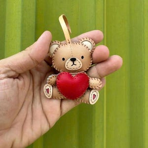May include: A small, tan teddy bear ornament with a red heart. The bear has stitched details, black eyes, and a beige loop for hanging. The heart is centered on the bear's chest. The background is a solid green.