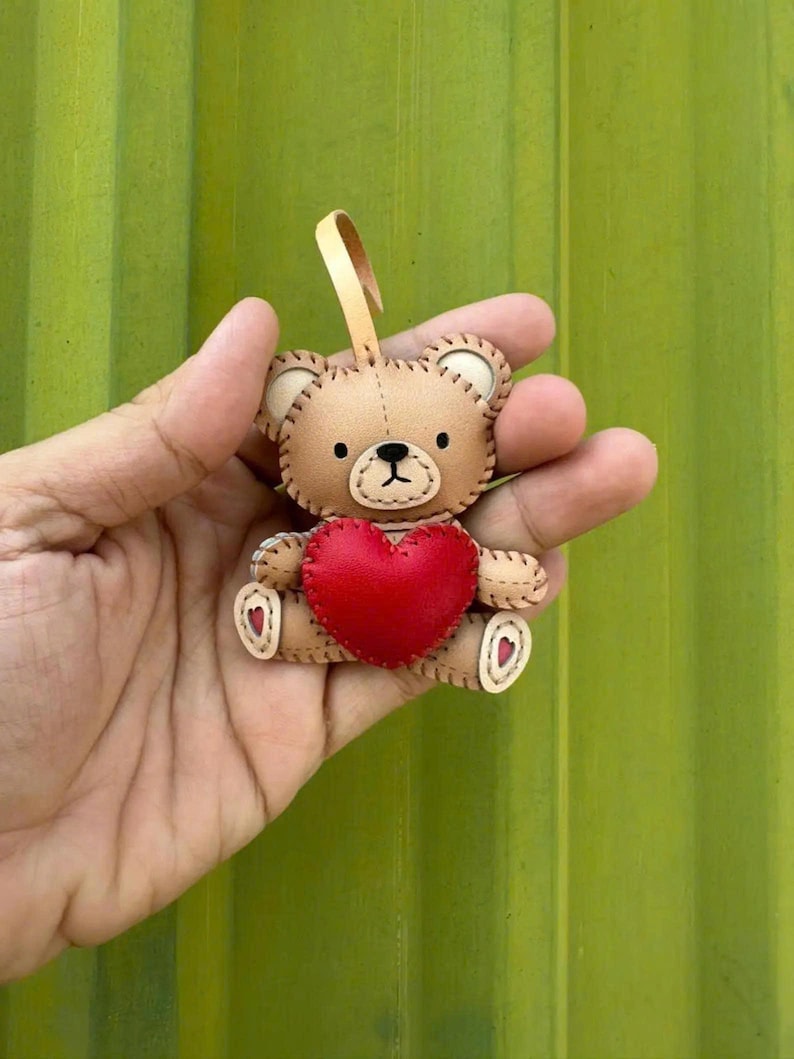 May include: A small, tan teddy bear ornament with a red heart. The bear has stitched details, black eyes, and a beige loop for hanging. The heart is centered on the bear's chest. The background is a solid green.