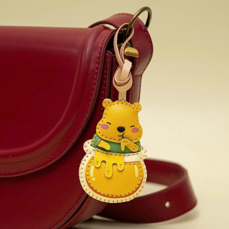May include: A deep red leather shoulder bag with a dangling charm shaped like a yellow bear character wearing a green shirt and holding a honey pot. The charm is attached by a light beige leather strap to a bronze-colored metal ring on the bag.
