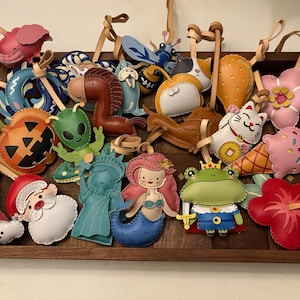 May include: A collection of colorful leather ornaments featuring various shapes including a flamingo, pumpkin, alien, mermaid, frog prince, Santa Claus, and flowers, each with a tan leather loop for hanging, displayed in a rectangular wooden tray.