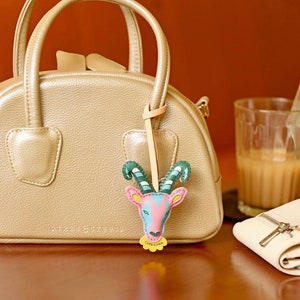 Puede incluir: Un bolso de mano de cuero beige con una cremallera dorada y un peque&ntilde;o charm de cuero colorido sujeto al asa. El charm tiene forma de cabra y lleva la palabra "Capricornio" escrita.