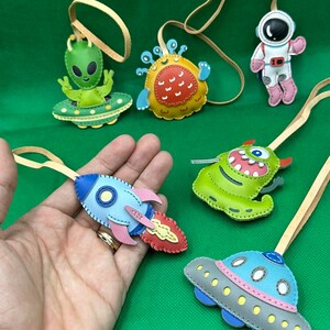 以下が含まれることがあります： 手作りのカラフルな革製宇宙をテーマにしたオーナメントのコレクション。オーナメントには、宇宙船に乗ったエイリアン、ロケット、モンスター、宇宙飛行士、UFOが含まれています。各オーナメントには、吊り下げ用のタン革ループが付いています。
