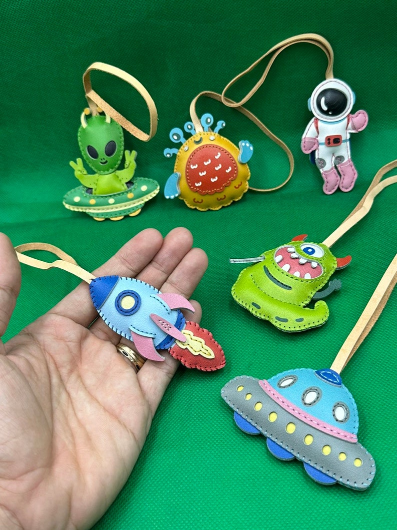 以下が含まれることがあります： 手作りのカラフルな革製宇宙をテーマにしたオーナメントのコレクション。オーナメントには、宇宙船に乗ったエイリアン、ロケット、モンスター、宇宙飛行士、UFOが含まれています。各オーナメントには、吊り下げ用のタン革ループが付いています。