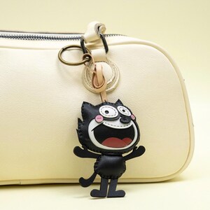 May include: A black cat keychain with a wide open mouth and surprised expression. The keychain is attached to a cream-colored leather bag with a gold-colored ring. The cat has white eyes, pink cheeks, and a red mouth.