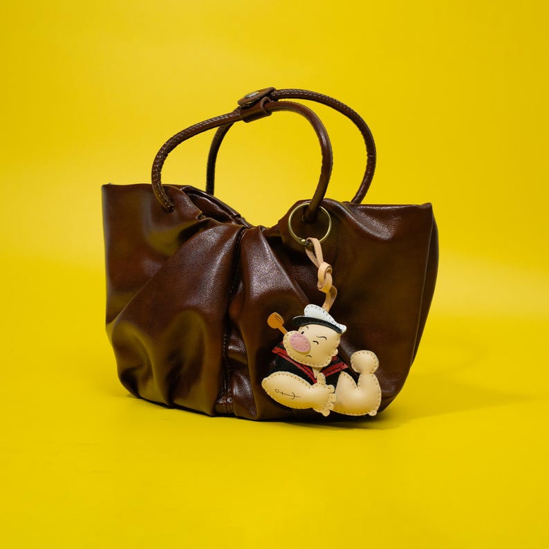 May include: A brown leather tote bag with a circular handle and a decorative charm. The charm is a cartoon character in a sailor hat. The bag is on a yellow background.