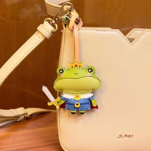 May include: A whimsical frog-shaped keychain with a crown and sword, attached to a cream-colored purse. The frog is green, blue, and red, adding a playful touch. The purse has a gold clasp and the word "JUNO" is visible.