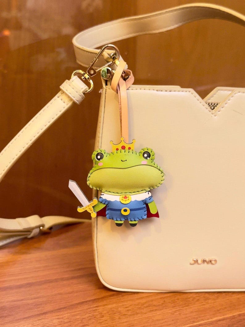 May include: A whimsical frog-shaped keychain with a crown and sword, attached to a cream-colored purse. The frog is green, blue, and red, adding a playful touch. The purse has a gold clasp and the word "JUNO" is visible.