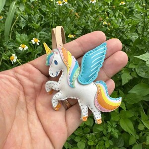 May include: A white unicorn ornament with a rainbow mane and tail, blue wings, and a yellow horn. The unicorn is held in a hand against a background of green foliage and small white flowers. A tan leather strap is attached to the top.