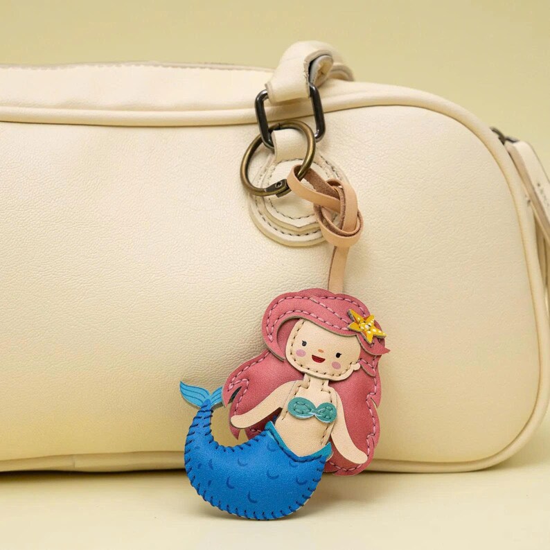 May include: A whimsical mermaid keychain with pink hair, a blue tail, and a gold starfish. The keychain is attached to a cream-colored purse with a leather strap and metal hardware. The mermaid is made of stitched leather.