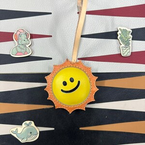 May include: A yellow leather sun with a smiling face, with orange stitching around the edges. The sun is hanging from a tan leather strap. The sun is on a background of black, white, and brown stripes.