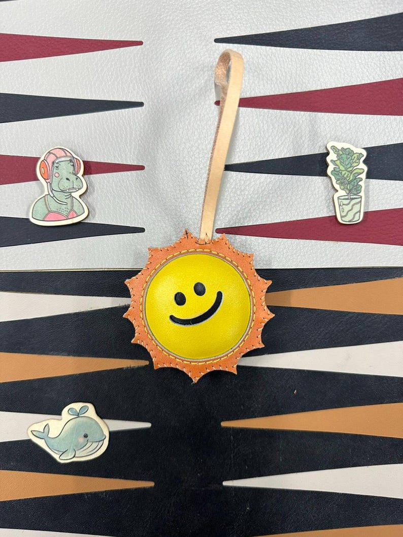 May include: A yellow leather sun with a smiling face, with orange stitching around the edges. The sun is hanging from a tan leather strap. The sun is on a background of black, white, and brown stripes.
