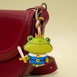 May include: A dark red leather handbag with a hanging frog-shaped charm. The charm depicts a green frog wearing a gold crown with red dots, a blue tunic with a red cape, and holding a white sword with a yellow hilt, attached by a light tan strap.