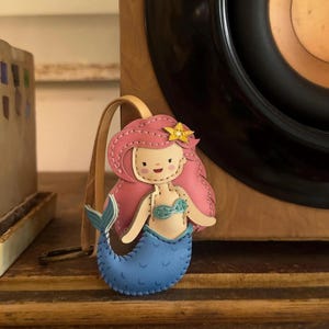 May include: A handmade mermaid keychain with pink hair, a yellow star, and a blue tail. The keychain is made of leather and has a tan strap. The mermaid is wearing a blue bikini top. The keychain is a cute accessory.