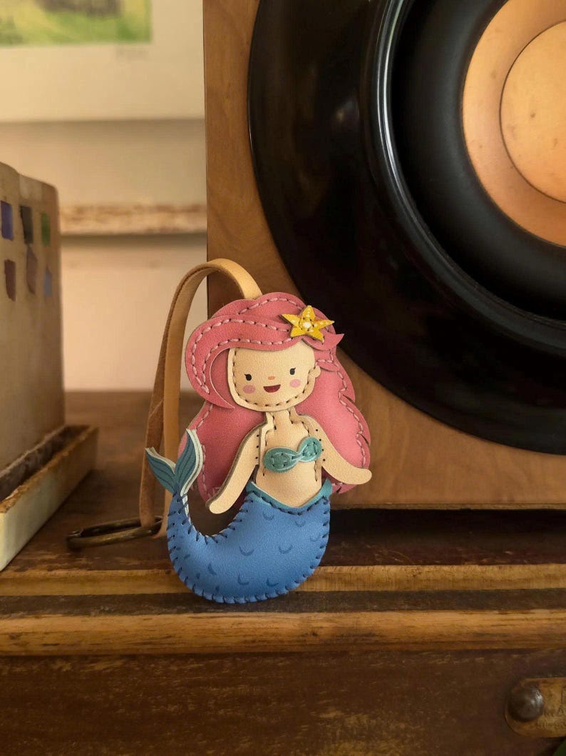 May include: A handmade mermaid keychain with pink hair, a yellow star, and a blue tail. The keychain is made of leather and has a tan strap. The mermaid is wearing a blue bikini top. The keychain is a cute accessory.