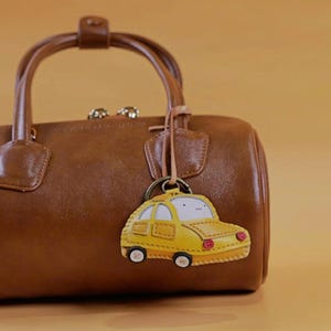 May include: A yellow taxi cab keychain with a smiling face and black wheels hangs from a brown leather handbag. The keychain has red headlights and a small tag. The handbag is a dark brown color.