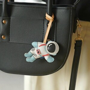May include: A black leather handbag with a small astronaut-shaped keychain attached. The keychain is white, blue, and red, with a detailed astronaut design. The bag has a top handle and a shoulder strap.