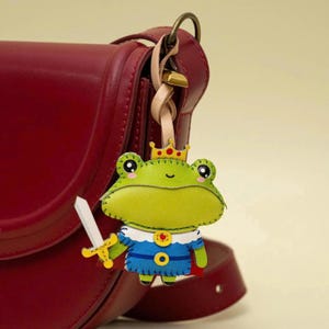 May include: A whimsical frog king keychain, crafted from leather, is attached to a red purse. The frog is green and yellow, wearing a blue outfit, a gold crown, and holding a sword. The keychain has a leather strap and gold-colored hardware.