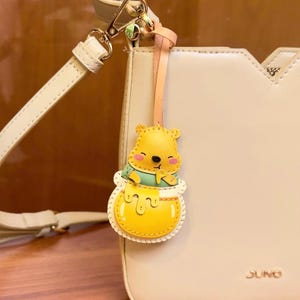 May include: A cream-colored handbag with a detachable shoulder strap and a decorative Winnie the Pooh charm. The charm features a yellow bear in a honey pot, with a light pink and peach-colored strap.