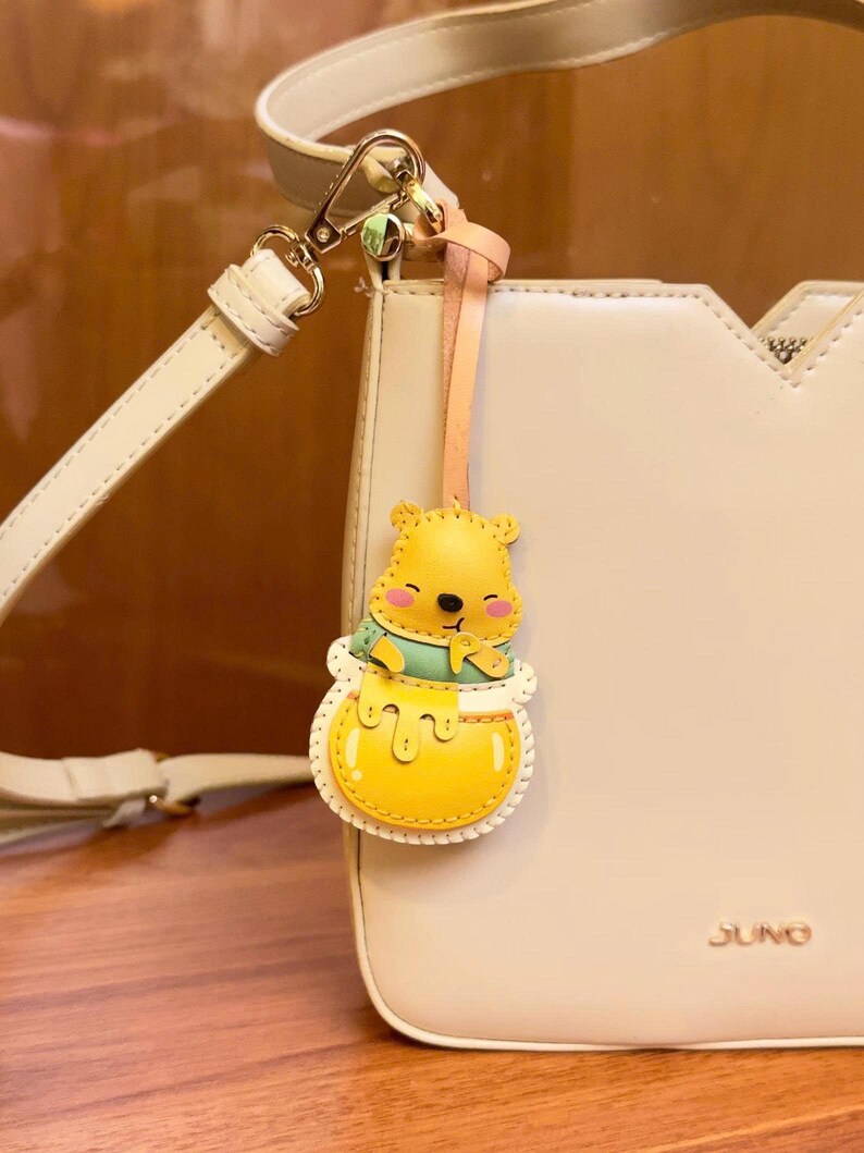 May include: A cream-colored handbag with a detachable shoulder strap and a decorative Winnie the Pooh charm. The charm features a yellow bear in a honey pot, with a light pink and peach-colored strap.