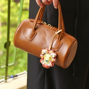Puede incluir: Un bolso de mano de cuero marr&oacute;n con una cremallera dorada y un dije de le&oacute;n. El dije tiene un dise&ntilde;o amarillo, naranja y verde con la palabra "LEO" impresa en &eacute;l.
