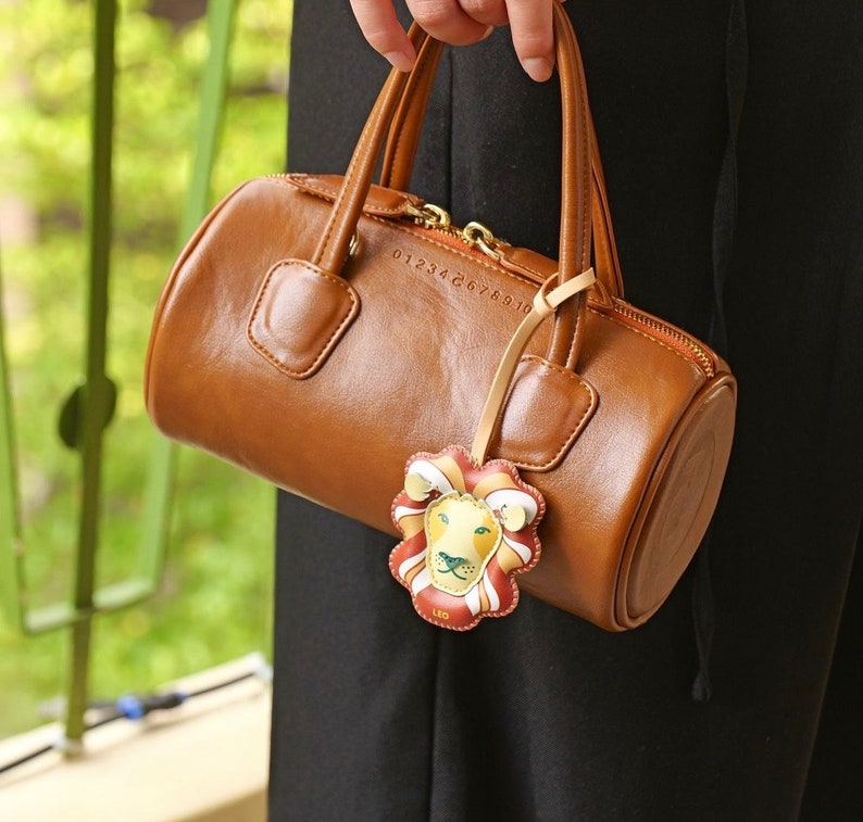 Puede incluir: Un bolso de mano de cuero marr&oacute;n con una cremallera dorada y un dije de le&oacute;n. El dije tiene un dise&ntilde;o amarillo, naranja y verde con la palabra "LEO" impresa en &eacute;l.