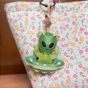 May include: A green leather alien keychain featuring a smiling extraterrestrial figure making a peace sign while sitting in a flying saucer with yellow polka dots, attached to a light-colored leather strap and a circular metal ring.