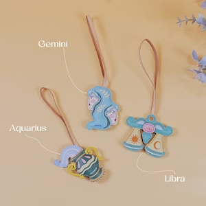 Puede incluir: Tres llaveros de cuero con signos del zod&iacute;aco: G&eacute;minis, Acuario y Libra. Cada llavero presenta un s&iacute;mbolo del zod&iacute;aco diferente en azul, amarillo y verde. Los llaveros cuelgan de una cuerda marr&oacute;n claro.