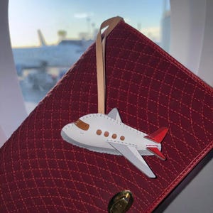 May include: A white leather airplane-shaped luggage tag with brown windows and red tail accents, attached to a tan leather strap. The tag rests against a quilted, deep red rectangular bag featuring a gold-toned metal turn-lock closure.