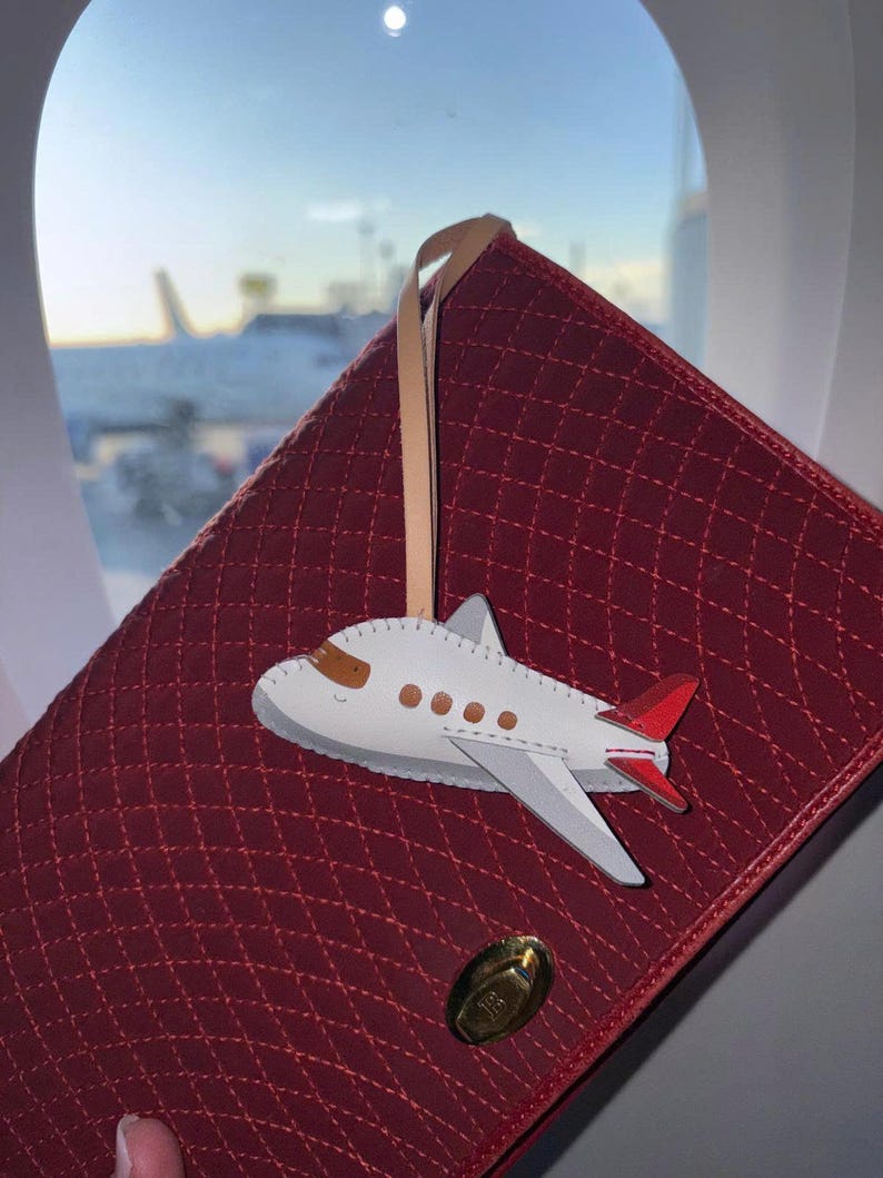 May include: A white leather airplane-shaped luggage tag with brown windows and red tail accents, attached to a tan leather strap. The tag rests against a quilted, deep red rectangular bag featuring a gold-toned metal turn-lock closure.