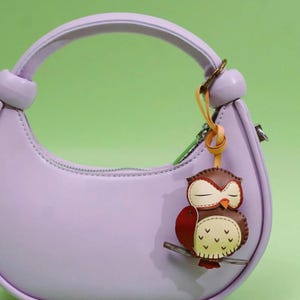 May include: A light purple handbag with a matching handle and a decorative owl keychain. The owl keychain is brown, red, and cream-colored, with a branch and a yellow leather strap. The background is a solid green.