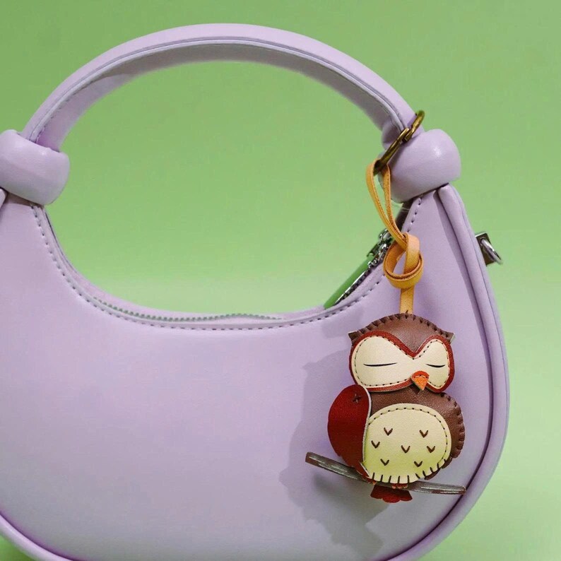 May include: A light purple handbag with a matching handle and a decorative owl keychain. The owl keychain is brown, red, and cream-colored, with a branch and a yellow leather strap. The background is a solid green.