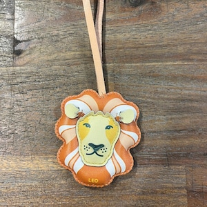 Puede incluir: Un llavero de cuero con un dise&ntilde;o de cara de le&oacute;n. La cara del le&oacute;n es naranja, amarilla y blanca. El llavero tiene una correa de cuero marr&oacute;n y la palabra "LEO" est&aacute; impresa en la parte inferior.