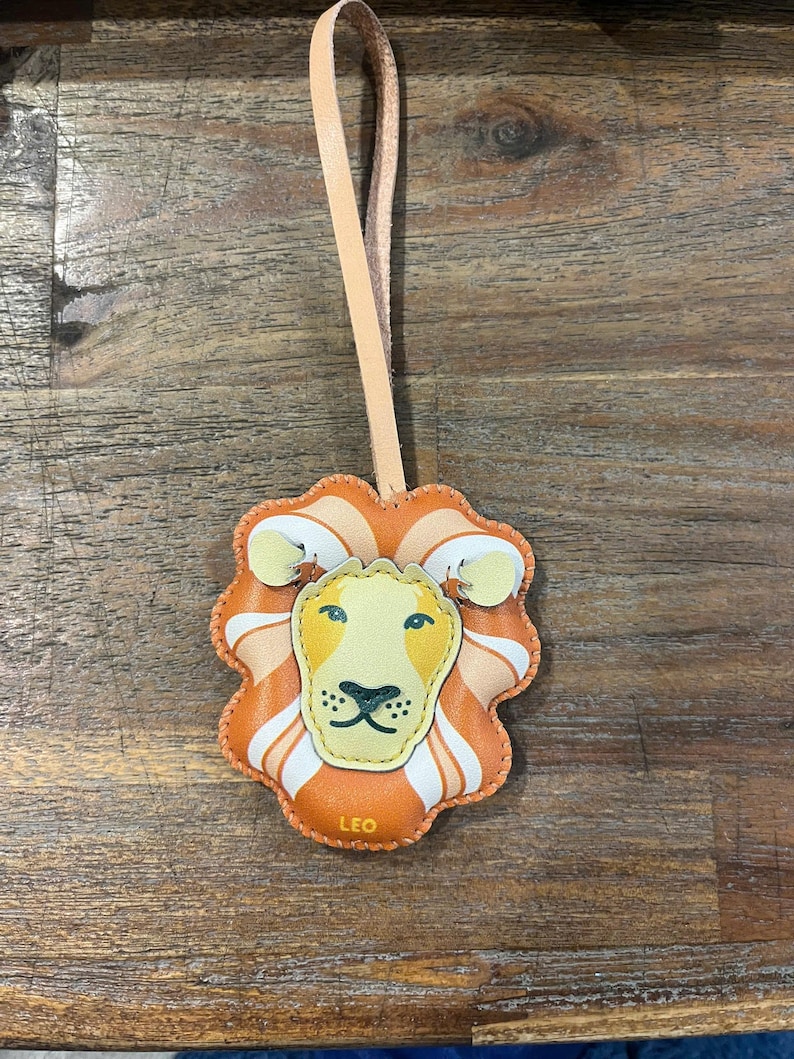 Puede incluir: Un llavero de cuero con un dise&ntilde;o de cara de le&oacute;n. La cara del le&oacute;n es naranja, amarilla y blanca. El llavero tiene una correa de cuero marr&oacute;n y la palabra "LEO" est&aacute; impresa en la parte inferior.