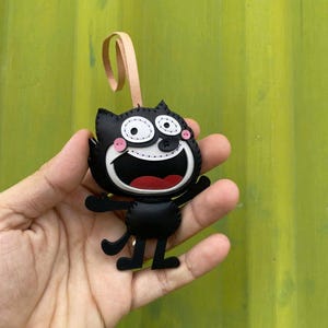May include: Black leather cat-shaped keychain with a smiling face, white eyes, and a red mouth. The keychain has a beige loop for hanging. The cat's arms are outstretched, and it has small black legs. The design is stitched with black thread.