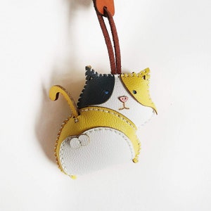 May include: A yellow, white, and black leather cat keychain with stitched details. The cat has a stitched face with blue eyes and a pink nose.