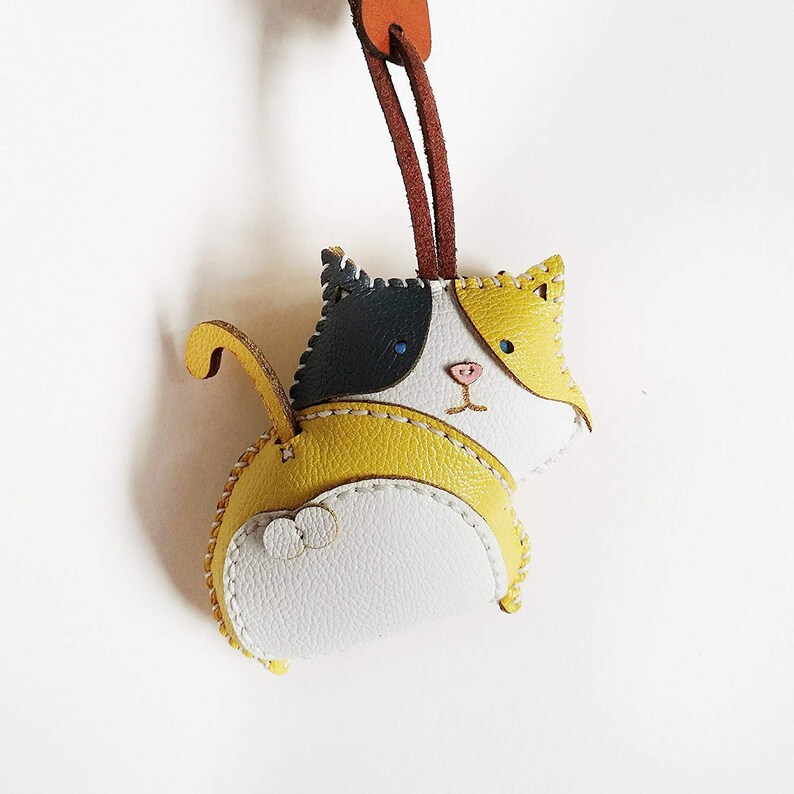May include: A yellow, white, and black leather cat keychain with stitched details. The cat has a stitched face with blue eyes and a pink nose.