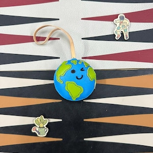 May include: A blue and green leather globe with a smiling face and a wink. The globe is hanging from a brown leather strap. The globe is on a black and white checkerboard background.