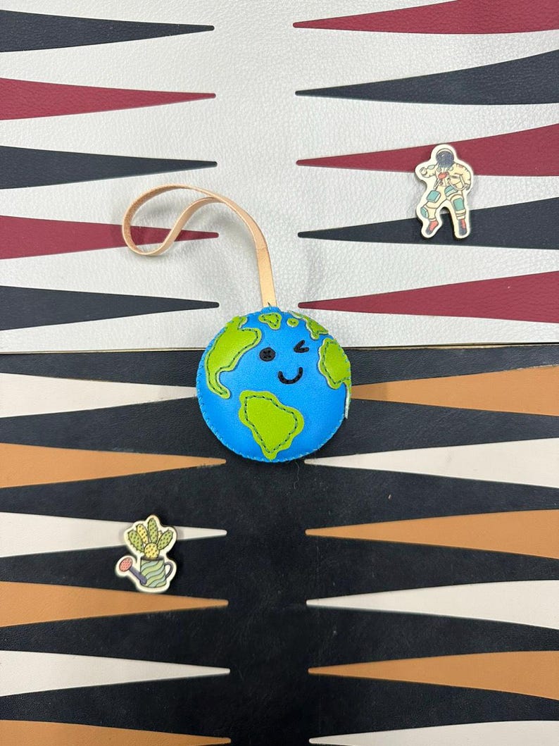 May include: A blue and green leather globe with a smiling face and a wink. The globe is hanging from a brown leather strap. The globe is on a black and white checkerboard background.