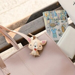 May include: A light pink leather tote bag with a matching strap, featuring a beige dog-shaped keychain with pink accents. A small illustrated notebook and a white wallet are also visible.