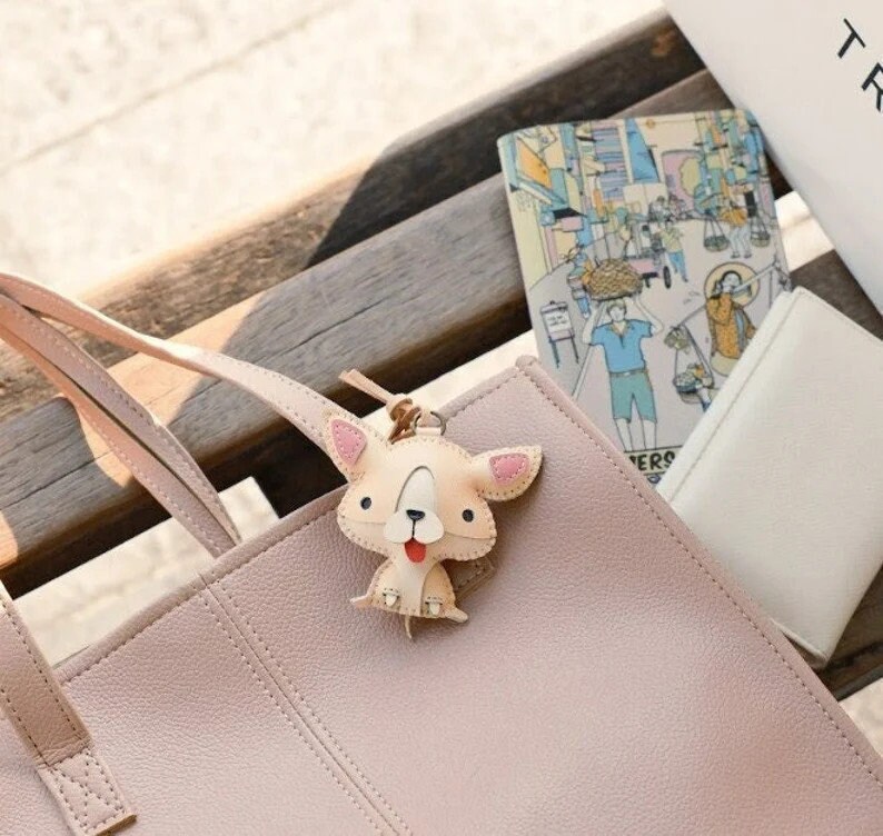 May include: A light pink leather tote bag with a matching strap, featuring a beige dog-shaped keychain with pink accents. A small illustrated notebook and a white wallet are also visible.