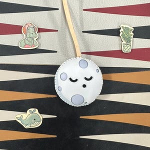 May include: A white felt moon with a stitched border and a sleeping face. The moon is hanging from a wooden dowel. There are three small wooden cutouts on a patterned background: a whale, a plant in a pot, and a person wearing headphones.