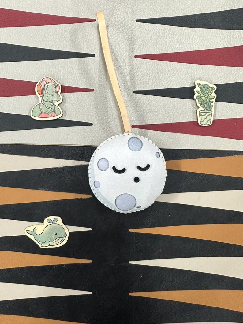 May include: A white felt moon with a stitched border and a sleeping face. The moon is hanging from a wooden dowel. There are three small wooden cutouts on a patterned background: a whale, a plant in a pot, and a person wearing headphones.