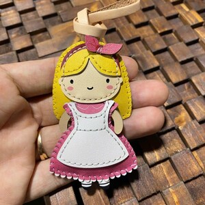 May include: A handmade leather keychain featuring a cartoon illustration of a girl with blonde hair, a pink bow, and a pink and white dress. The keychain has a tan leather loop for attachment.