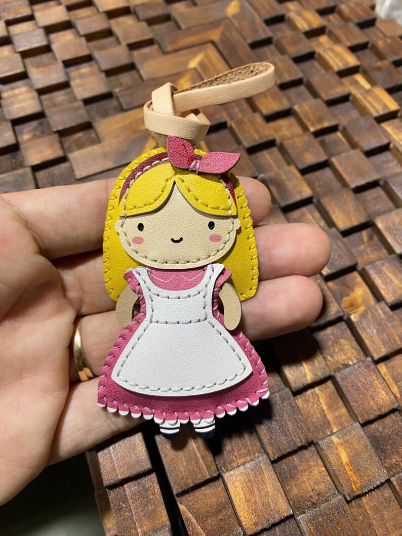 May include: A handmade leather keychain featuring a cartoon illustration of a girl with blonde hair, a pink bow, and a pink and white dress. The keychain has a tan leather loop for attachment.