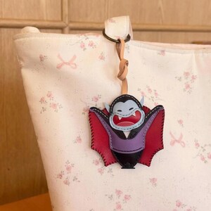 May include: A leather vampire-shaped keychain with a red and black cape, purple vest, and blue face. The keychain is attached to a light-colored bag with a floral pattern. The keychain is hanging from a leather strap.