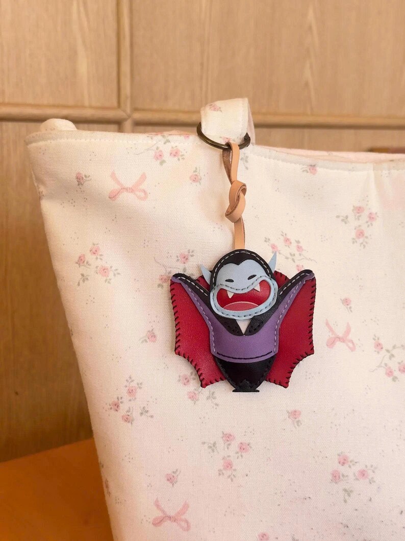 May include: A leather vampire-shaped keychain with a red and black cape, purple vest, and blue face. The keychain is attached to a light-colored bag with a floral pattern. The keychain is hanging from a leather strap.